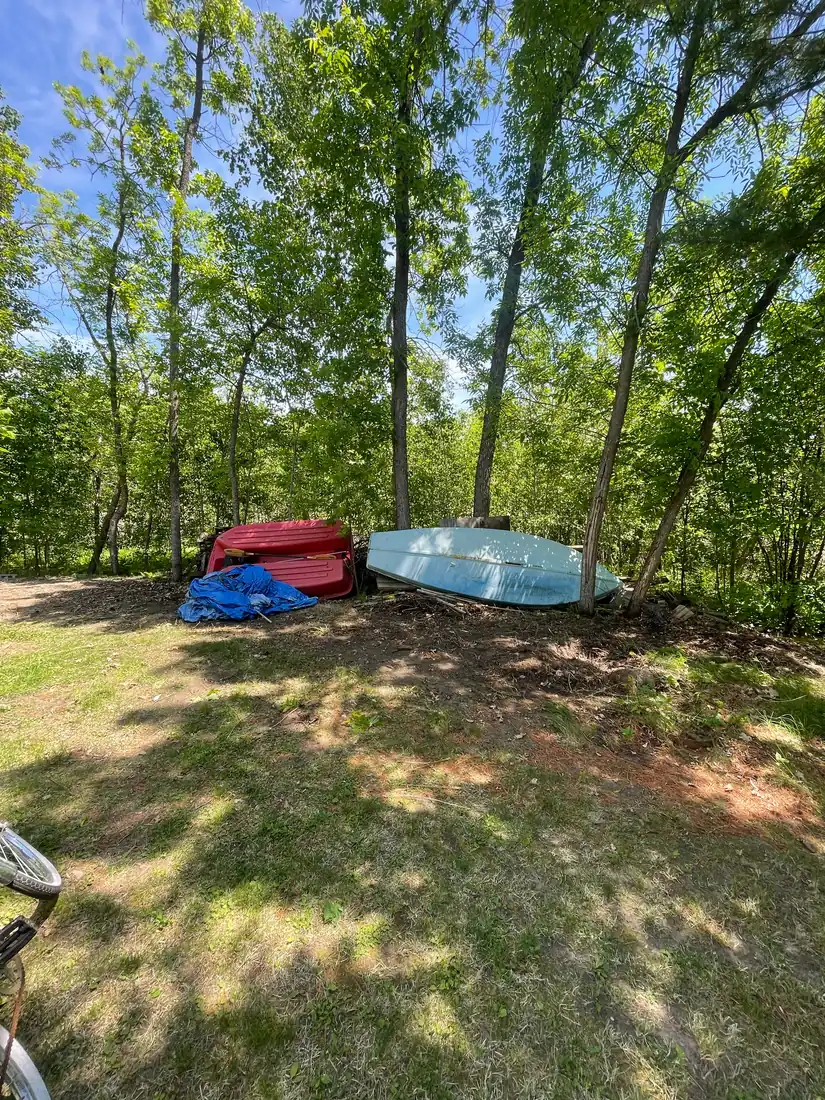 The same wooded lot edge with the brush pile cleared and the ground raked clean. The boat and truck bed were not part of the job and remain in place.