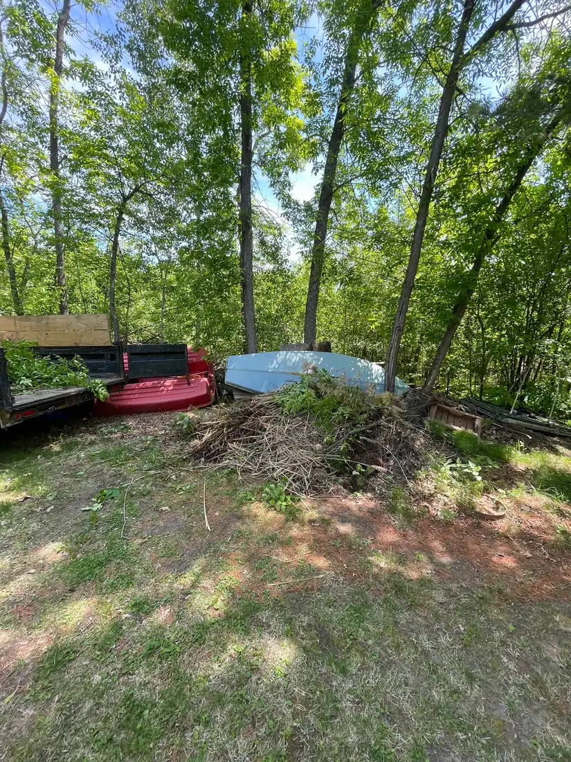 Large pile of cut brush and dead branches at the edge of a wooded lot in Itasca County, beside an overturned aluminum boat and an old truck bed.